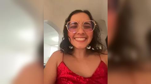 Media: Video of a smiling, fair-skinned woman with curly black hair, wearing large round glasses and a red sequined dress, taken from a slightly blurry frontal angle.