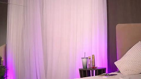 Media: Video of a minimalist bedroom with white curtains, a beige headboard, and a bedside table holding a glass, lotion, and remote control. The room is softly lit with purple lighting, creating a serene atmosphere.