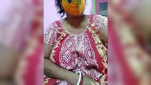 Media: Video of an Indian woman wearing a red and white patterned sari, with a yellow face mask covering her nose and mouth, seated with arms crossed. Background includes a light pink wall and blurred furniture.