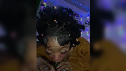 Media: Video of a young Black woman with curly hair, wearing a mustard-yellow sweater, lying on her stomach, looking down at her phone, surrounded by colorful fairy lights in a dimly lit room.