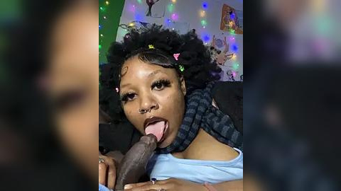 Media: Video of a Black woman with dark curly hair, wearing a nose ring, and licking an erect penis in a room adorned with colorful lights and posters.