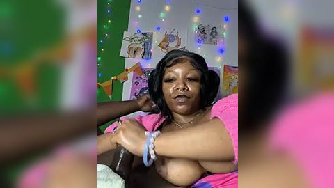 Media: A video shows a plus-sized Black woman with large breasts, wearing headphones, in a colorful, festive room with string lights, art, and a pink blanket.