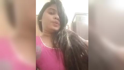 Media: A blurry video of a woman with long, dark hair, wearing a pink shirt, making a kissy face. Background features a white wall and a locker.