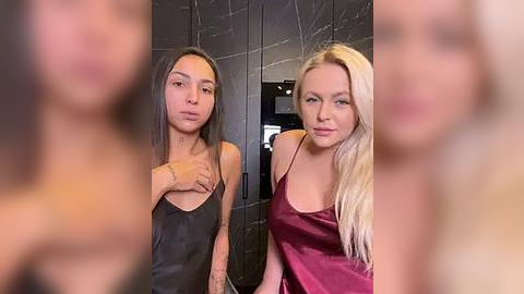 Media: Video of two women, one with long dark hair in a black tank top, the other with long blonde hair in a red satin dress, standing in a modern, darkly tiled bathroom.