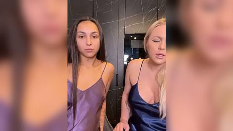 Media: Video of two women in dark, textured, black marble-walled room. One with long black hair in purple satin camisole, another with blonde hair in blue satin camisole.