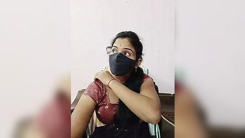 Media: Video of a young woman with dark hair, wearing a red-patterned blouse, black mask, and black headband, sitting indoors, hands on her chest, looking concerned.