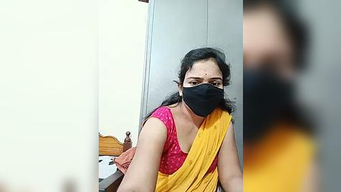 Media: A video of a woman with dark skin and black hair, wearing a pink polka-dotted blouse, a yellow sari, and a black mask, sitting on a wooden bed in a simple, sparsely decorated room.