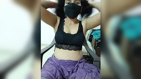 Media: Video of a woman with medium skin tone, wearing a black lace bralette and purple pants, sitting on a chair with a black face mask, holding her hair.