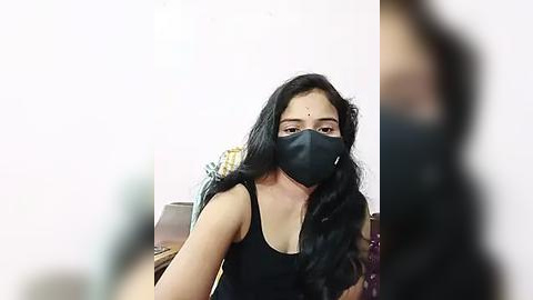 Media: Video of a South Asian woman with long black hair, wearing a black face mask and black tank top, seated at a desk in a dimly lit room.