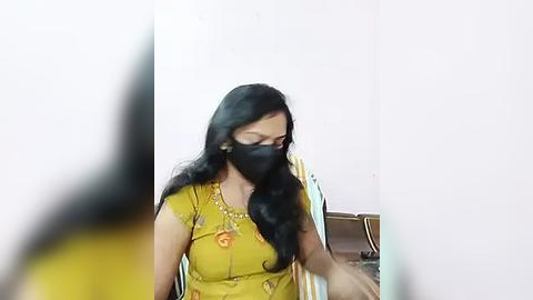 Media: Video of a woman with long black hair, wearing a yellow top with floral embroidery, a black face mask, and seated on a chair, blurred background.