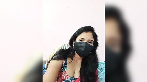 Media: Video of a South Asian woman with long black hair, wearing a floral blouse, black face mask, and a blue headscarf, blurred in the foreground.