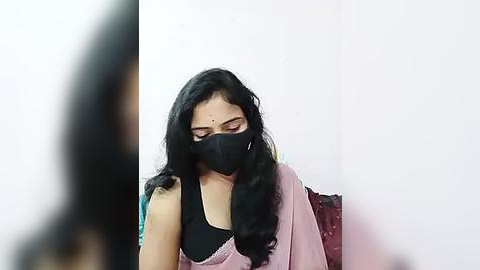 Media: Video of a South Asian woman with long black hair, wearing a black mask, pink saree, and black top, sitting in a white room.