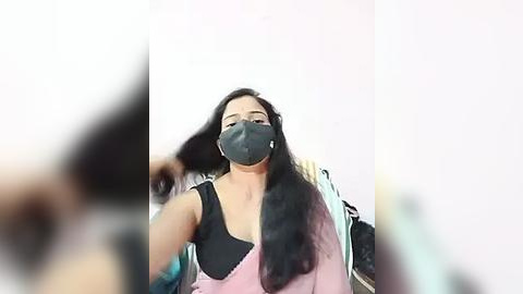 Media: A video of a woman with long black hair, wearing a gray face mask, black tank top, and pink jacket, adjusting her hair, taken from a low angle.