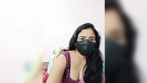 Media: Video of a South Asian woman with long black hair and medium skin tone, wearing a black mask, pink blouse, and a traditional Indian sari, sitting on a bed with a blurred background.