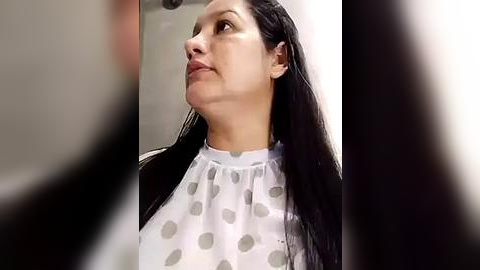 Media: Video of a woman with long black hair, wearing a white blouse with grey polka dots, standing indoors with a blurred background.