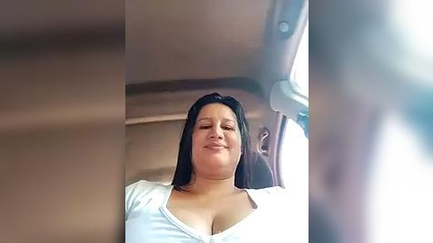 Media: Video of a smiling, light-skinned woman with shoulder-length black hair, wearing a white top, seated in a car. The background shows a blurred, beige interior with a window.