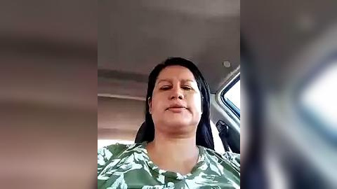 Media: Video of a middle-aged woman with medium skin tone, long black hair, and a light green floral-patterned blouse, driving a car, taken from the front passenger seat.