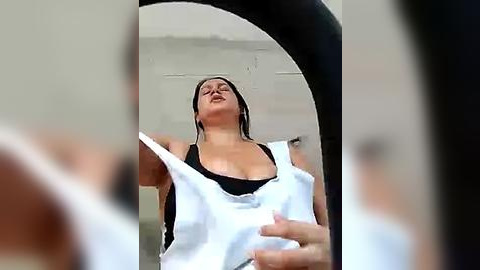 Media: Video of a woman with short, dark hair, wearing a black tank top, lifting a white shirt in front of a mirror, appearing to be in a bathroom with tiled walls.