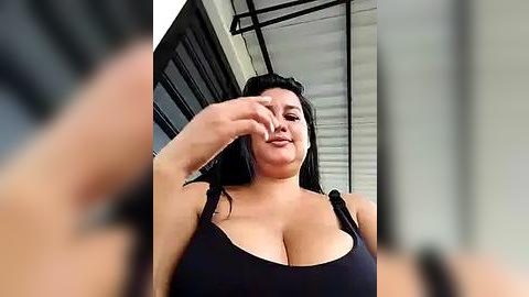 Media: A video of a plus-size woman with long black hair, wearing a low-cut black tank top, and a black arm covering her face. She stands in a modern, industrial-style room with metal beams and glass windows.