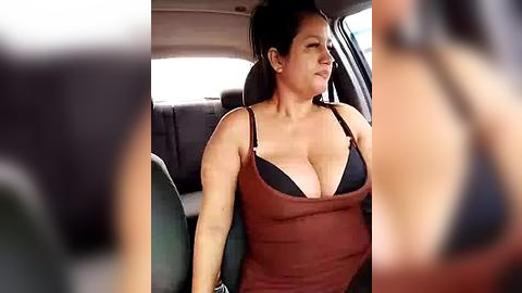 Media: Video of a middle-aged woman with fair skin, wearing a revealing brown tank top that exposes her black bra, sitting in a car seat.