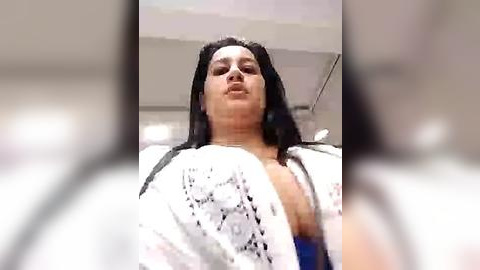 Media: Video of a plus-size woman with long black hair and a white lace cardigan, standing indoors. The image is slightly blurred, focusing on her upper body.