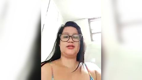 Media: A video of a plus-sized woman with long dark hair, wearing glasses and a colorful tank top, standing in a brightly lit room with a window.