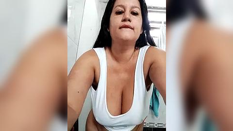 Media: A video of a middle-aged Latina woman with long black hair and fair skin, wearing a low-cut white tank top that reveals cleavage, standing in a modern bathroom.