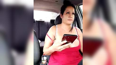 Media: Video of a Latina woman with medium skin tone and dark hair in a bun, wearing a pink tank top and holding a smartphone, seated in a car.