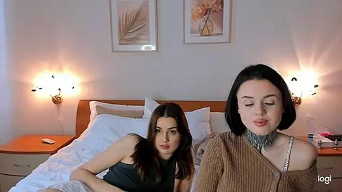 Media: Video of two young women in a modern bedroom: one with dark hair and a tattooed neck, the other with long dark hair, lying on a bed.