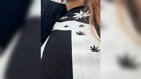 Media: Video of a woman's upper body wearing a black and white jacket with white cannabis leaf patches, visible in close-up. The background is blurred, focusing attention on the jacket's texture and design.