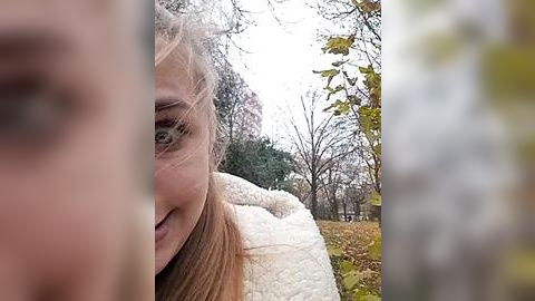 Media: Video of a young woman with fair skin and light brown hair, smiling, partially visible through a blurry, textured window. Background features autumn trees and a white, fluffy object, possibly a pillow.