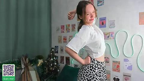 Media: Video of a young woman with light skin and brown hair in a white crop top and leopard print pants, posing in a festive room with a Christmas tree, wall decorations, and a neon sign.