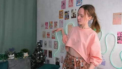 Media: Video of a young woman with light skin and brown hair, wearing a peach crop top and patterned skirt, surrounded by colorful anime posters and a lit-up Christmas tree in a cozy, eclectic room.