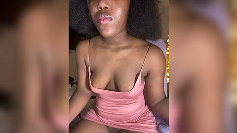 Media: Video of a Black woman with medium-dark skin, wearing a pink spaghetti-strap dress, exposing cleavage, sitting on a bed with a gray blanket. Her curly hair frames her face, and she has a relaxed, slightly pouting expression.