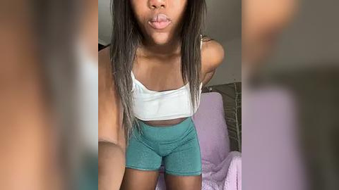 Media: Video of a Black woman with long, straight, dark hair, wearing a white crop top and teal shorts, playfully sticking out her tongue, indoors.