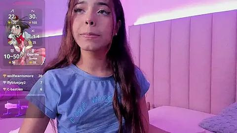 Media: Video of a young woman with long brown hair, wearing a blue t-shirt, standing in a room with a purple and pink lighting ambiance.