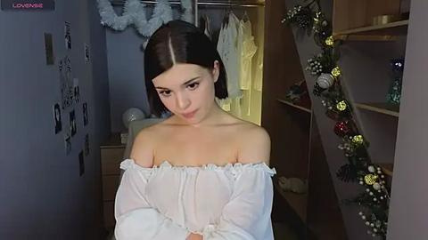 Media: Video of a young woman with straight dark hair, wearing an off-shoulder white dress, standing in a cozy, dimly lit room with Christmas decorations and a wardrobe visible in the background.