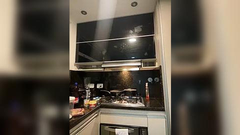 Media: Video of a modern, well-lit kitchen with a black countertop, stainless steel appliances, a black backsplash, and a large overhead range hood. The background is blurred.