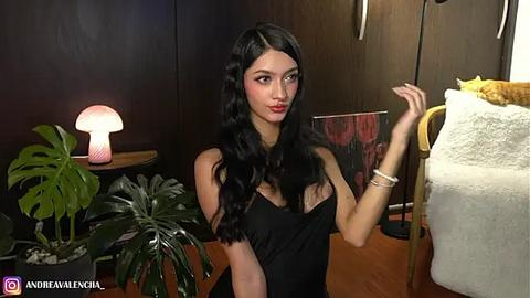 Media: Video of a slender, fair-skinned woman with long black hair, wearing a black dress, holding a cigarette, indoors with dark wooden walls, a lamp, potted plants, and a towel.