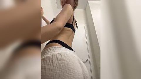 Media: Video of a young woman with fair skin, wearing a black bra and white shorts, standing in a narrow hallway with white walls and a silver door.