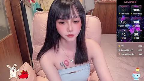 Media: Video of a young Asian woman with straight black hair, light skin, and a small tattoo on her chest, sitting on a beige couch in a cozy room, with an overlay showing livestream stats.