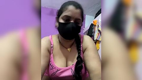 Media: Video of a young woman with medium skin tone and dark hair, wearing a pink tank top, black face mask, and gold necklace, in a dimly lit room with purple walls.