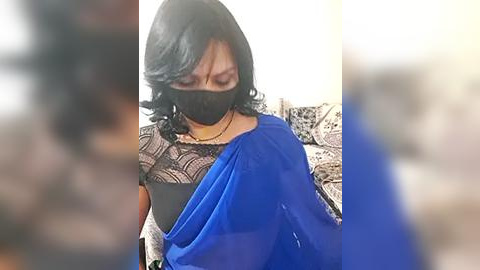 Media: Video of a woman with black hair wearing a black face mask, blue saree, and black blouse, sitting on a couch with patterned cushions in a dimly lit room.