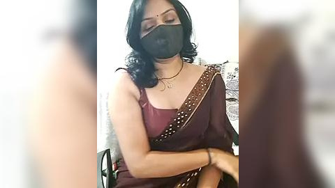 Media: Video of a South Asian woman with fair skin, dark hair, wearing a maroon saree with gold embroidery, black face mask, and a gold necklace.