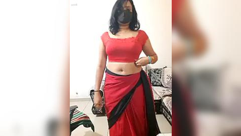 Media: Video of a woman in a red and black saree, black mask, and black shoes, standing in a living room with a couch and colorful clothes on the floor.