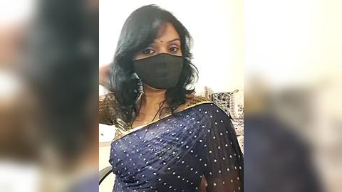 Media: Video of an Indian woman in a blue sari with white polka dots, wearing a black mask, standing indoors with blurred background.