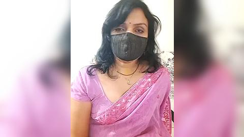 Media: Video of an Indian woman with medium skin tone, wearing a black face mask, a pink sari with intricate embroidery, and a black top. Background is blurred.