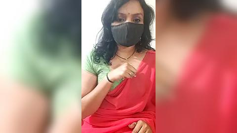 Media: Video of an Indian woman in a red sari with a green blouse, wearing a black face mask, with blurred green and red figures in the background.