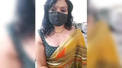 Media: Video of a South Asian woman with medium brown skin, wearing a black face mask, a green and yellow sari with a white pattern, and a black blouse with a white design. She has shoulder-length black hair and is indoors.