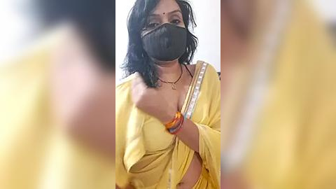 Media: Video of a woman in a yellow sari with a black face mask, wearing a gold necklace and bangles. Background is blurred, focusing on her from the waist up.